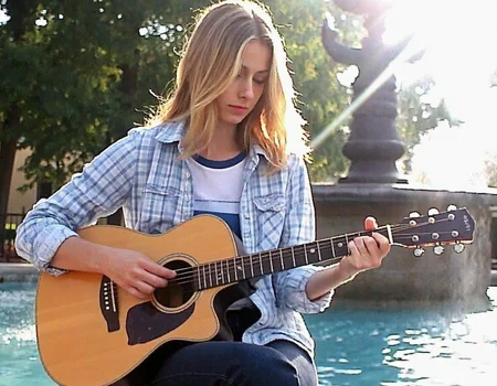 v8s, a college woman with freckles is sitting outside on the ledge of a fountain in a park playing the guitar, bright lighting casts lens flares, she has shoulder-length blond straight hair and is wearing a light blue plaid shirt with rolled up sleeves to her elbows, revealing a plain white t-shirt with a blue collar underneath, as well as dark blue jeans. Amateur quality, Analog distortion, low resolution, Chromatic aberration, tape noise, Grainy texture, Frame jitter, muted colors, overexposed foreground