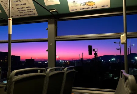 Bus interior, empty seats visible, gray and patterned fabric, metal handrail with white plastic handle. Large windows with a vibrant sunset, vivid pink and purple hues, contrasting the city skyline silhouette. Glimpses of buildings, streetlights, and distant industrial structures visible outside. Upper panel features a diagram with Japanese text, depicting a hand holding a card, advertisements in Japanese, muted colors. No people present, focus on the serene contrast between interior and exterior. Wide-angle lens, good quality image., 
