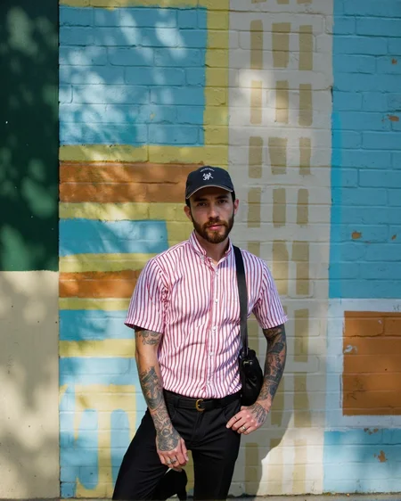 A man with a beard and tattoos standing in front of a colorful striped wall, posing confidently with one hand on his hip and the other resting on his thigh. he is wearing a red and white striped button-up shirt, black pants, and a black bag slung over his shoulder. his body is slim, and he has a beard. he has several tattoos on his arms, including one on his left arm and another on his right arm. he also has a cap on his head and a ring on his finger. the man is looking directly at the camera with a serious expression. the background is a vibrant outdoor setting with a striped wall in shades of blue, green, and yellow.