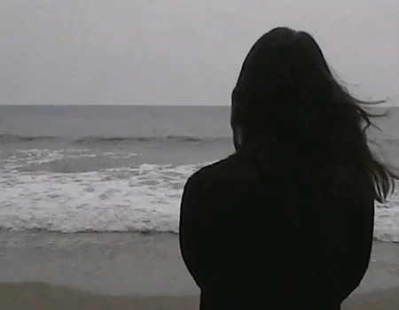 v8s, woman, from back view, dark silhouette, dark long hair, wearing dark shirt, standing on beach, ocean waves, horizon line, overcast sky, underexposed lighting