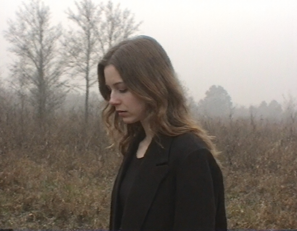 v8s, Young woman, mid-20s, light brown wavy hair, fair skin, wearing a dark oversized coat, standing in profile with head slightly bowed, somber expression, no visible makeup, natural look, scene set in an outdoor marshland, visible reeds and bare trees, overcast sky, mist in the background, muted earthy tones, introspective mood, low lighting, eerie sky and atmosphere