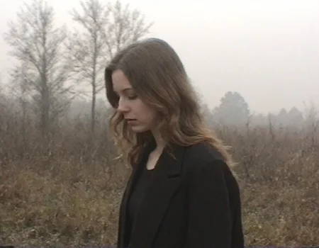 v8s, Young woman, mid-20s, light brown wavy hair, fair skin, wearing a dark oversized coat, standing in profile with head slightly bowed, somber expression, no visible makeup, natural look, scene set in an outdoor marshland, visible reeds and bare trees, overcast sky, mist in the background, muted earthy tones, introspective mood, low lighting, eerie sky and atmosphere