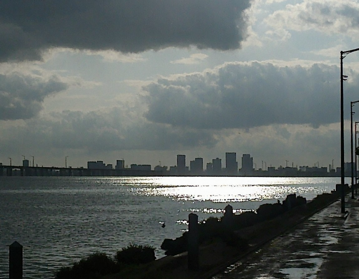 v8s, Overcast sky with dense clouds, sun peeking through, strong reflections on water, wide river or body of water, distant bridge spanning water, modern city skyline silhouette, high-rise buildings in background, empty road alongside water, tall streetlights lining road, wet pavement suggesting recent rain, early morning or late afternoon light, hazy atmosphere, dim lighting, outdoor setting, high contrast between bright reflections and dark clouds