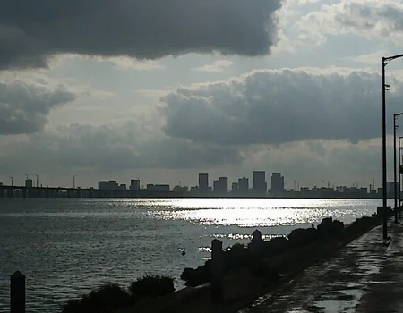 v8s, Overcast sky with dense clouds, sun peeking through, strong reflections on water, wide river or body of water, distant bridge spanning water, modern city skyline silhouette, high-rise buildings in background, empty road alongside water, tall streetlights lining road, wet pavement suggesting recent rain, early morning or late afternoon light, hazy atmosphere, dim lighting, outdoor setting, high contrast between bright reflections and dark clouds