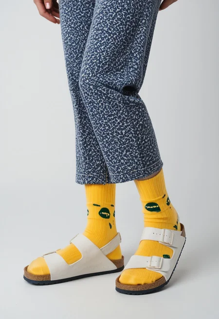 (photograph of woman's feet wearing socks and sandals and leggings), 1girl, solo, dynamic_pose, simple_background, white_background, scksnsndls, patterned blue and yellow socks, lower_body, footwear_focus, ankles, calves, birksonsocks, birkenstock