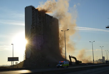 Demolition of high-rise building, partially collapsed structure, cranes on site, hydraulic excavators visible, dense rising dust cloud, early morning or late afternoon, warm sunlight backlighting structure, roadway in foreground, silhouetted street lamps, distant cityscape background, billboard with indistinct advertisement, ambulance driving on road, clear sky with few clouds, several contrails visible, urban setting, heavy machinery in action, dramatic sunburst effect through debris, bokeh background, natural lighting, overexposed highlights., 