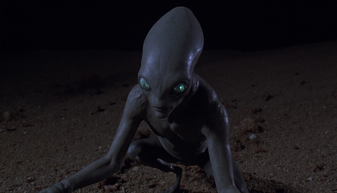 cinematic close-up shot of a grey alien crouching in the desert at night. Cold harsh artificial light coming from above as a cone and reflecting off its skin. High resolution. 1990s movie <lora:dark_cinematic-000080:1>