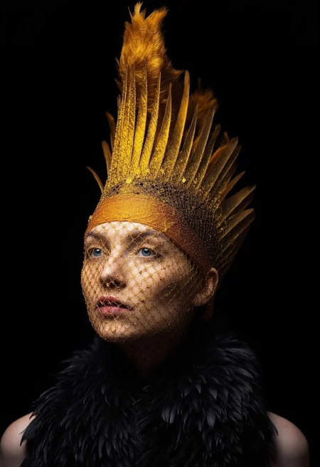 A surreal portrait of a woman with a striking, almost otherworldly appearance. She is captured in a three-quarter view, with her face and shoulders prominently displayed against a pitch-black background. The lighting in the image is dramatic, with a warm, golden glow emanating from the left side of her face, casting soft shadows and highlighting her features. Her skin is pale, and her facial features are finely sculpted with sharp, defined contours. She has dark, wavy hair that is pulled back, allowing the focus to remain on her face. Her eyes are a striking, intense blue, and she wears an elaborate, golden veil that covers her entire face except for her lips, which are painted a muted pink. The veil is intricately designed with a delicate, feather-like texture that radiates outward from her face, creating a dramatic and ethereal effect. The feathers are a mix of gold and bronze, adding a luxurious and almost regal aura to her appearance. She is adorned with a high collar made of dark, fluffy feathers, which adds to the exotic and fantastical nature of the image. The textures in the feathers are meticulously rendered, with each individual feather's details visible, enhancing the realism of the artwork. The overall composition is both elegant and mysterious, evoking a sense of ancient mythology or fantasy.