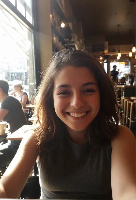 A casual iPhone selfie of a 24-year-old woman sitting in a coffee shop, taken at armâs length. She has a playful smile, slightly squinting as sunlight hits her face from the window. The blurred café background shows people chatting, baristas making coffee, and soft warm lighting.