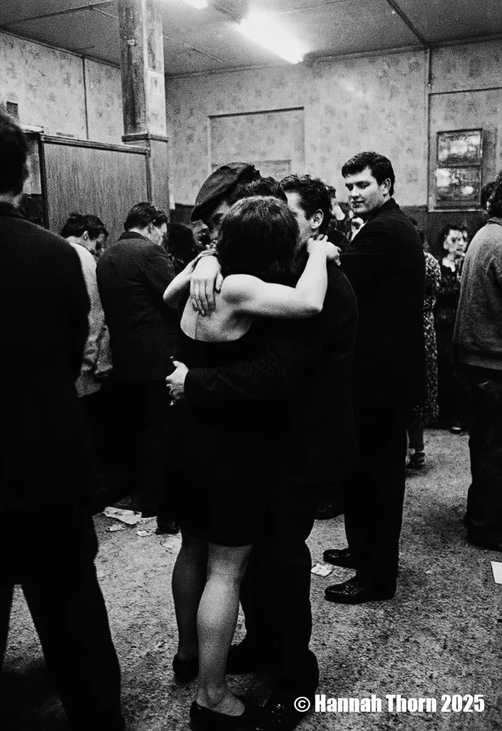 A black and white photograph from a low angle, depicting a scene of a couple embracing in a crowded room filled with people. the couple, a man and a woman, appear to be in their mid-twenties, with the woman wearing a short black dress and the man wearing a cap. they are standing close together, their faces close together as they hug each other tenderly. the woman's hands are on the man's waist, and they are facing away from the camera. in the background, several other men and women are standing, some of them looking at the couple. the room is dimly lit, with a few framed pictures on the wall, and there are papers scattered on the floor. the overall mood of the image is one of intimacy and closeness, as if the couple is in the midst of a romantic moment.<lora:_Flux\Anders_Petersen.safetensors:0.9:0.9>
