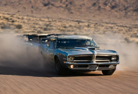 Create an ultra-detailed and realistic image of a race between 2 custom cars, created with parts from many muscle cars, with aerodynamic lines from 1972. The cars have tinted black windows. It is captured in motion with motion blur at full speed through a desert, daylight. Race between 2 cars. The photograph is captured with an Arri Alexa camera, with a zoom lens. <lora:Vintage Muscle Car SDXL:1>