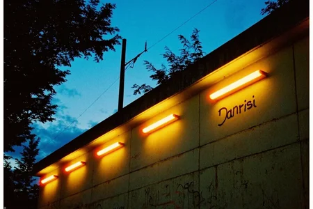 8n8log, Concrete wall, industrial structure, row of five orange fluorescent lights mounted along top edge, casting warm yellow glow against pale green-tinted concrete blocks, Stylish graffiti tag "Danrisi" visible on wall, silhouettes of trees visible above wall, electrical power lines and utility pole visible on left side, twilight blue sky in background, dusk setting, urban or industrial environment, slight film grain texture, low exposure in shadow areas, cool-warm color contrast between blue sky and orange lights, outdoor setting, evening time