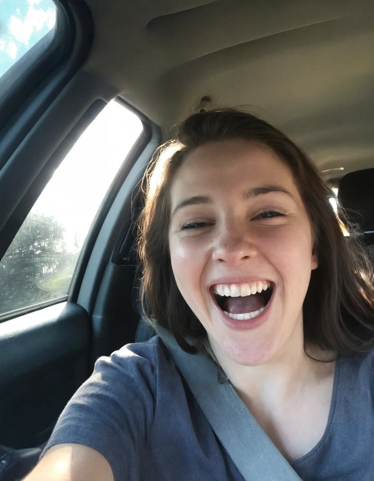 Authentic share, Spontaneous, authentic, natural light: Car selfie. Person in driver's seat, light coming through windshield. Seatbelt visible, dashboard and steering wheel in frame. Subject possibly caught mid-laugh or making a face.