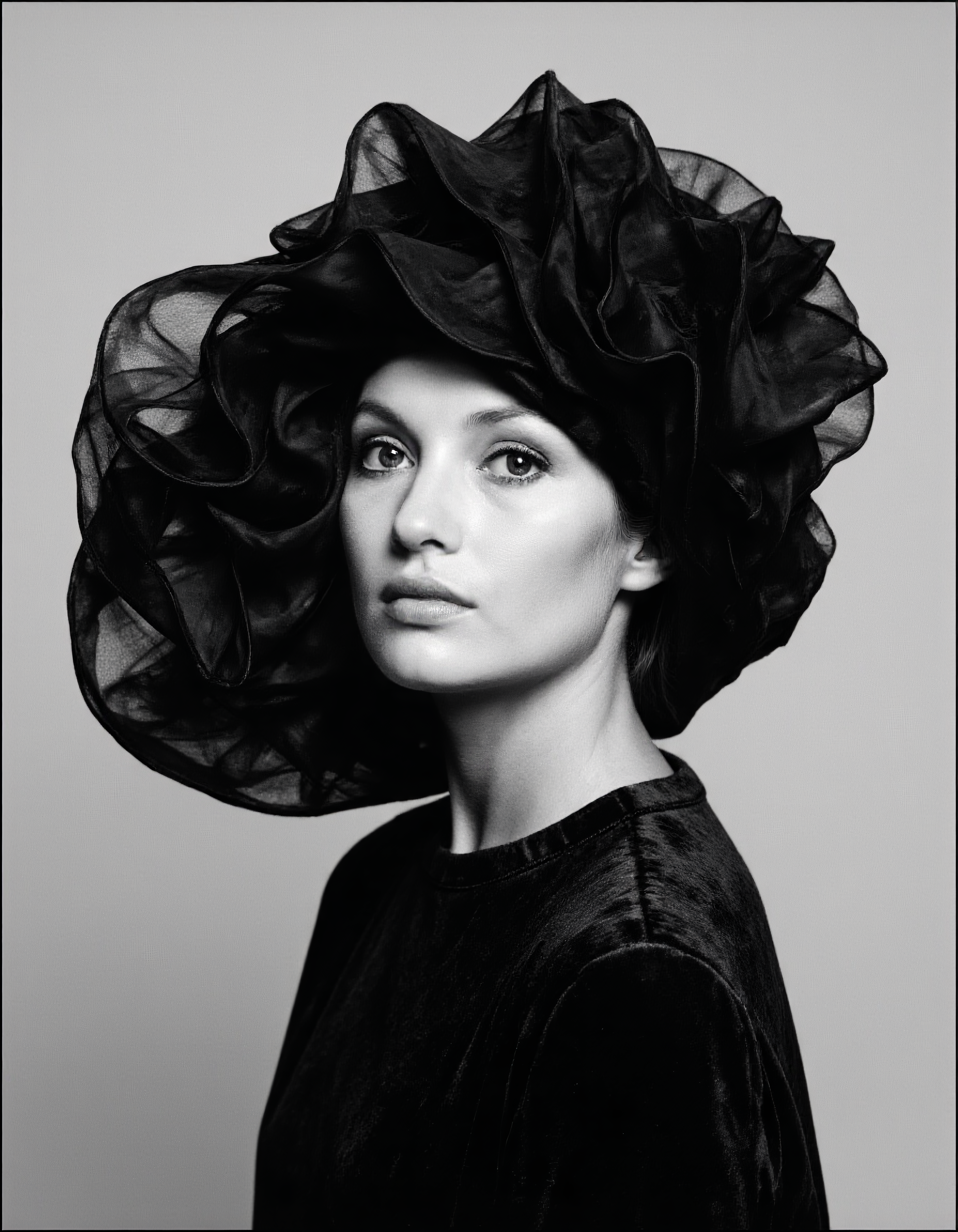 A high-contrast photo of a woman posing for a picture, captured in a dramatic black and white portrait as she wears an avant-garde, oversized hat that partially obscures her face. Evoking the distinctive style of David Bailey, this image exude drama and mystery, reminiscent of iconic mid-20th century fashion photography. The background minimalistic and slightly blurred to keep the focus on the subject, with subtle shadows and highlights enhancing the depth and dimension. The woman position slightly off-center to the left, with her hat extending towards the right, following the rule of thirds and aligning her eyes with the upper third of the image. A monochromatic palette with deep blacks and bright whites, and soft, directional lighting from the upper left to cast dramatic shadows on the right side of her face and hat. The hat features intricate, wavy textures for movement and depth, and sharply define her facial features, particularly her eyes and lips. The fabric of her clothing have a velvety texture for added luxury. The image with a medium-format camera at eye level for an intimate and detailed shot. <lora:david-bailey-flux1-dev-v1:1>