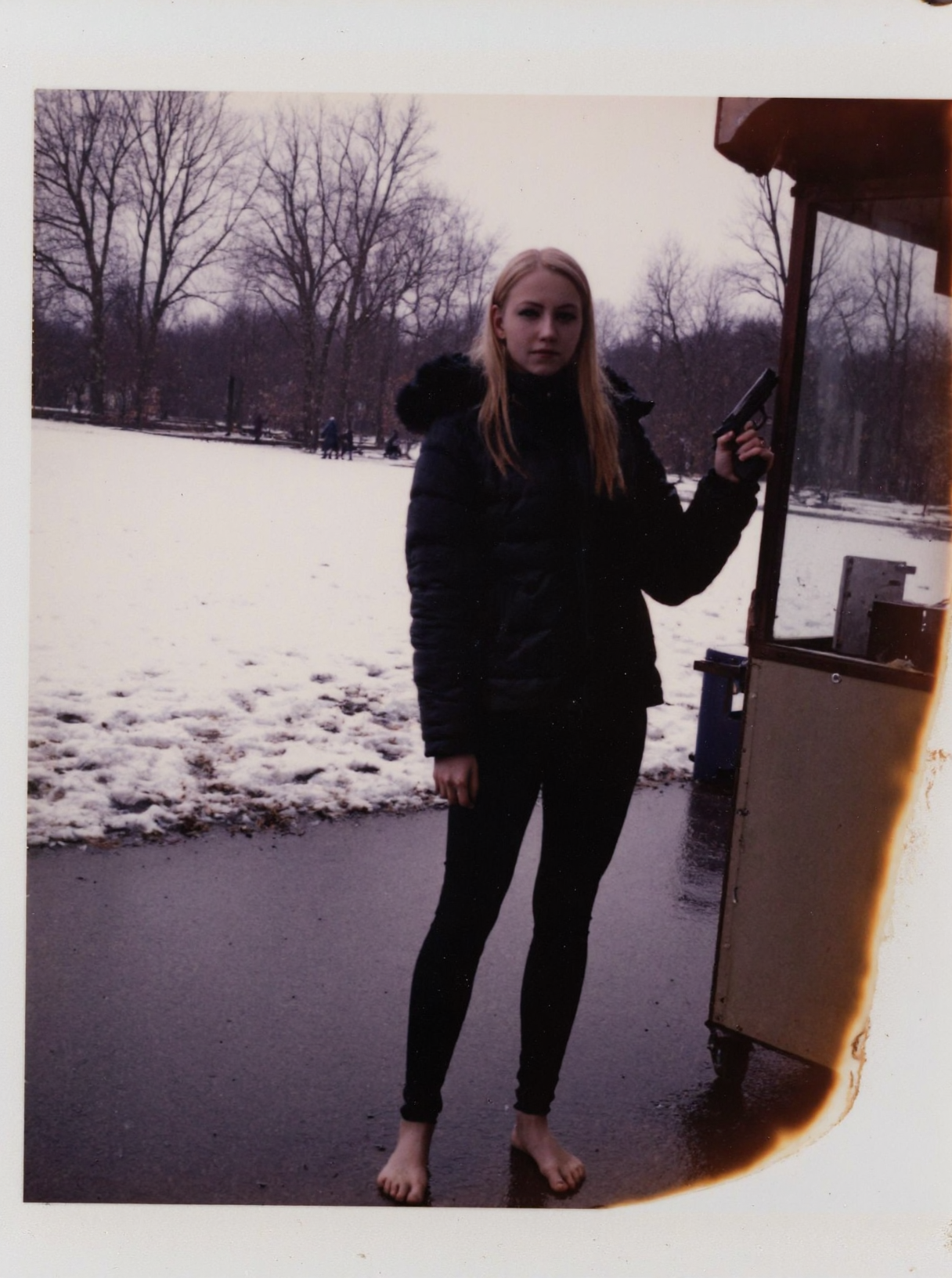 p0l2rd, Young woman, early 20s, casual winter attire, wearing a black puffer jacket with a faux fur hood, tight black pvc leggings, no shoes, barefoot on wet snowy ground. She stands at a food stall outdoors, holding a handgun right hand, left hand gesturing at viewer. Blonde long ponytail, natural makeup. Background shows a park, snow-covered ground, footprints, overcast weather, trees in distance, wintery atmosphere.  prominent film grain, high contrast lighting casting dramatic shadows, grainy film-like texture,  polaroid-style format with white border, distinctive burnt edge, photograph appears aged or partially developed, with the right side fading into white
