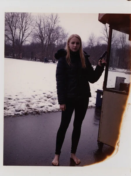 p0l2rd, Young woman, early 20s, casual winter attire, wearing a black puffer jacket with a faux fur hood, tight black pvc leggings, no shoes, barefoot on wet snowy ground. She stands at a food stall outdoors, holding a handgun right hand, left hand gesturing at viewer. Blonde long ponytail, natural makeup. Background shows a park, snow-covered ground, footprints, overcast weather, trees in distance, wintery atmosphere.  prominent film grain, high contrast lighting casting dramatic shadows, grainy film-like texture,  polaroid-style format with white border, distinctive burnt edge, photograph appears aged or partially developed, with the right side fading into white