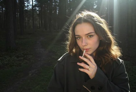 low-resolution photo, shot on a mobile phone, noticeable graininess, no visible JPEG artifacts, some lens flare from the sunlight in the background. Young woman, early 20s, casual style, wearing dark trench coat, long wavy brown hair cascading over her shoulders, looking direct at the viewer, makeup, outdoors in the dark forest, autumn, holding a cigarette to her lips, smoking cigarette, sunlight filtering through the trees