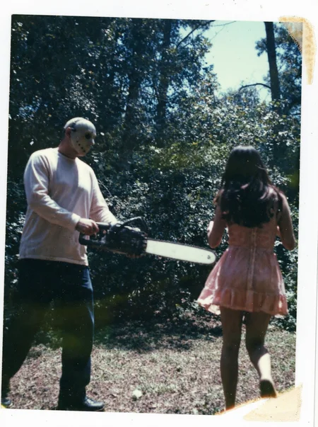 p0l2rd, Overexposed and color-distorted instant photo, man wielding a running chainsaw, facing towards a young woman running away, tall trees and dense foliage filling the background, man wearing a light-colored long-sleeve shirt, dark pants, dark shoes, and scary hockey mask. The woman with long dark hair, a light pink or coral dress, and dark shorts, the sunlight through the trees creates lens flares and hazy patches, outdoor forest setting, daytime, likely a staged or posed scene, damaged edges, colors shifted toward blue and green tones. prominent film grain, high contrast lighting casting dramatic shadows, grainy film-like texture