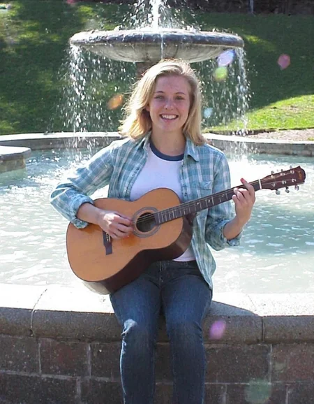 m8vic2, college woman with freckles is sitting outside on the ledge of a fountain in a park playing the guitar, bright lighting casts lens flares, she has shoulder-length blond straight hair and is wearing a light blue plaid shirt with rolled up sleeves to her elbows, revealing a plain white t-shirt with a blue collar underneath, as well as dark blue jeans