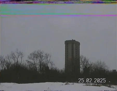 v8s, A still image with vhs effect, a tall building in the distance, bare trees in the foreground, a cloudy sky, timestamp 25.02.2025 in the lower right corner, snow on the ground, a horizontal band of static noise at the top of the image, a dark color palette, a sense of desolation.