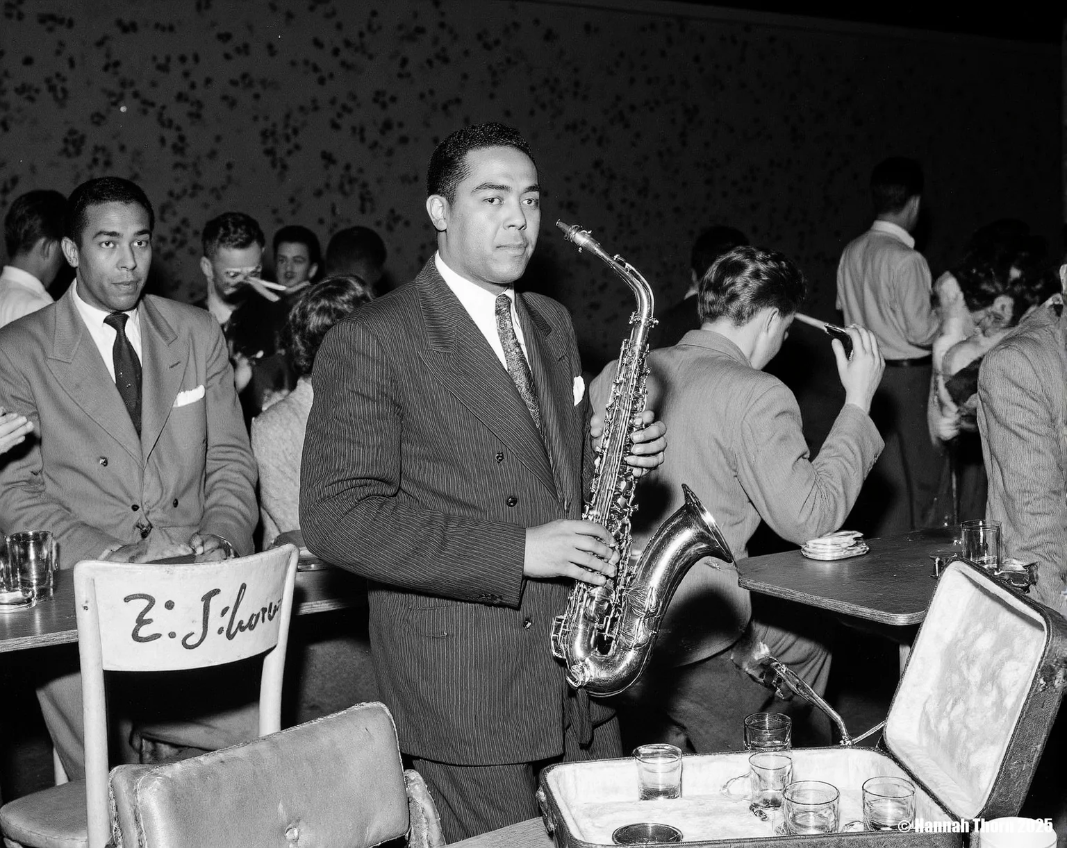 The image is a black and white photograph of the renowned jazz saxophonist, Lester Young, seemingly at a club or bar.    **Description:**  Lester Young is centrally positioned, holding a saxophone. He's dressed in a dark suit and tie, projecting a sophisticated yet relaxed demeanor. The saxophone is partially nestled in its case, which is open and rests on a table. The table is also occupied by what appears to be small glasses or cups and an ashtray.   Behind and to the side of Young are several people, some appear to be patrons of the establishment. One woman is visible, smoking a cigarette; others are partially obscured.  A sign that reads "E.E. Charles" is on the back of a chair behind the table.  The background shows a dark, possibly dimly lit interior with some indistinct wall markings or decorations.  The overall atmosphere appears to be that of a lively yet intimate setting.   **Style:**  The style of the photograph is classic black and white photojournalism or candid portraiture.  The contrast is strong, typical of the era, likely produced from film negatives. The grain is visible, adding to its documentary feel. The composition is fairly straightforward, drawing the viewer's attention immediately to Lester Young. The candid nature of the shot, with other patrons present, makes it seem like a spontaneous capture of a moment rather than a formally posed portrait. The grainy texture and the high contrast between light and shadow are characteristic of mid-20th-century street photography and jazz photography styles.  The slightly blurred background also helps to focus the viewer's eye on Lester Young and his saxophone. <lora:_Flux\Vintage_Jazz-Club.safetensors:1.0:1.0>