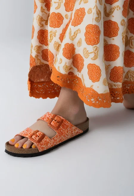 1girl, solo, simple_background, white_background, birksonfeet, orange_toenail_polish, close-up, lower_body, footwear_focus, patterned, long_skirt