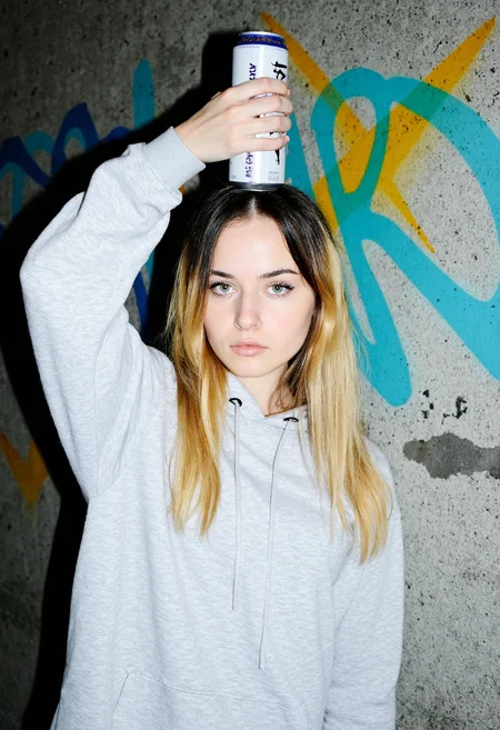 Young woman, early 20s, wearing a light grey oversized hoodie, straight posture, balancing a tall can of energy drink on top of her head with a steady left hand, pale complexion, long hair parted in the middle with dark roots transitioning to light blonde, subtle makeup with emphasis on eyes, neutral lips, indoors, standing against a heavily graffitied concrete wall in vibrant colors, overexposed, amateurish quality., 