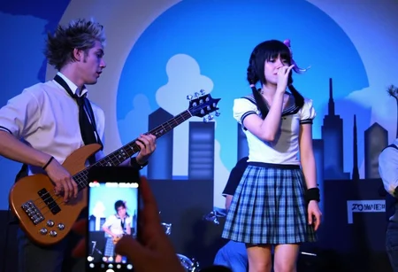 This is a photograph capturing a live music performance. The image features two musicians and a female vocalist on stage, set against a backdrop resembling a city skyline. The backdrop includes a large, blue, crescent moon and skyscrapers. The woman on the right is dressed in a white, short-sleeve blouse and a plaid skirt, reminiscent of a school uniform. She has her hair styled in pigtails, accessorized with a purple hair accessory. Her right arm is raised, holding a microphone close to her mouth, indicating she is singing. She wears a black wristband on her left arm. The male musician on the left plays an electric bass guitar, with a white shirt and black tie, his hair styled in a messy, spiky fashion. He is focused on his instrument, playing with intensity.In the foreground, a person's hand holds a flip phone, possibly recording the performance. The lighting is dim, typical of an indoor concert venue, with the stage illuminated by stage lights. The overall atmosphere suggests an energetic, youthful vibe, possibly a punk or rock concert