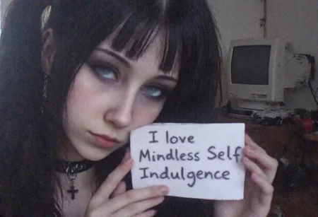 v8s, Young woman, early 20s, slim build, light skin, dark straight hair styled in pigtails, blunt bangs covering forehead, wearing black choker and black cross necklace, slightly smirk, three-quarters angle, looking directly at the viewer, head slightly leaned, left hand holding a crumpled piece of paper with handwritten text "I love Mindless Self Indulgence", dressed in casual dark-toned clothes, standing indoors in a low-lit room, background showing cluttered furniture, desk with retro computer and crt monitor, muted and desaturated colors