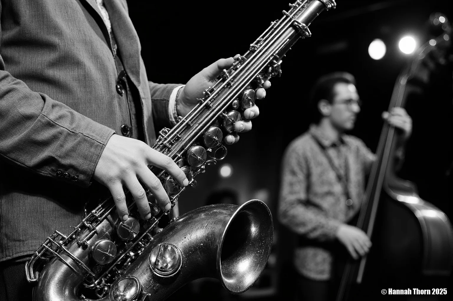 The image is a black and white close-up shot of a saxophonist playing a tenor saxophone. The focus is sharply on the saxophone and the musician's hands, which are positioned on the instrument's keys and valves. The saxophone itself appears aged, with a slightly worn look to its metallic surface. The details of the instrument, such as its keys and bell, are clearly visible. The saxophonist is wearing a somewhat loose-fitting jacket and a visible portion of a collared shirt underneath. The background is significantly out of focus, but shows a blurry figure, seemingly another musician playing an upright bass, suggesting a jazz or blues performance setting. The background lighting is dim, with some blurred light sources visible, contributing to the moody atmosphere. The style of the photograph is distinctly **black and white photojournalism** or **concert photography**. The high contrast, shallow depth of field (emphasizing the saxophone while blurring the background), and the candid feel (as if capturing a moment during a live performance) are all typical of these styles. The monochrome palette enhances the dramatic effect and focuses attention on the texture and form of the saxophone and the musician’s interaction with it. The overall mood is intimate and intense, conveying the emotion and energy of a live music performance. <lora:_Flux\Vintage_Jazz-Club.safetensors:1.0:1.0>