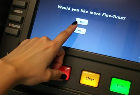 Hand of woman with black nailpolish about to press button, pale skin, ATM screen displaying message "Would you like more Fine-Tune?", options Yes and No, finger pointing at button in front of Yes, grey plastic buttons. Cancel button in red, Clear button in yellow, Enter button in green, outdoor setting, low lighting, screen glow providing illumination, slight glare on screen, high angle view