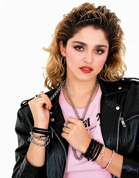 Madonna a woman posing for a picture against a white background. She has a confident and determined expression on her face, her hair is styled in side ponytail goals and her makeup with dark eyebrown, dark eyeliner and mascara and red lipstick. She is wearing black leather jacket, pink t-shirt, 80's bracelet, 80s earrings, several different types of 80s neck chains and her hands are clasped in front of her.