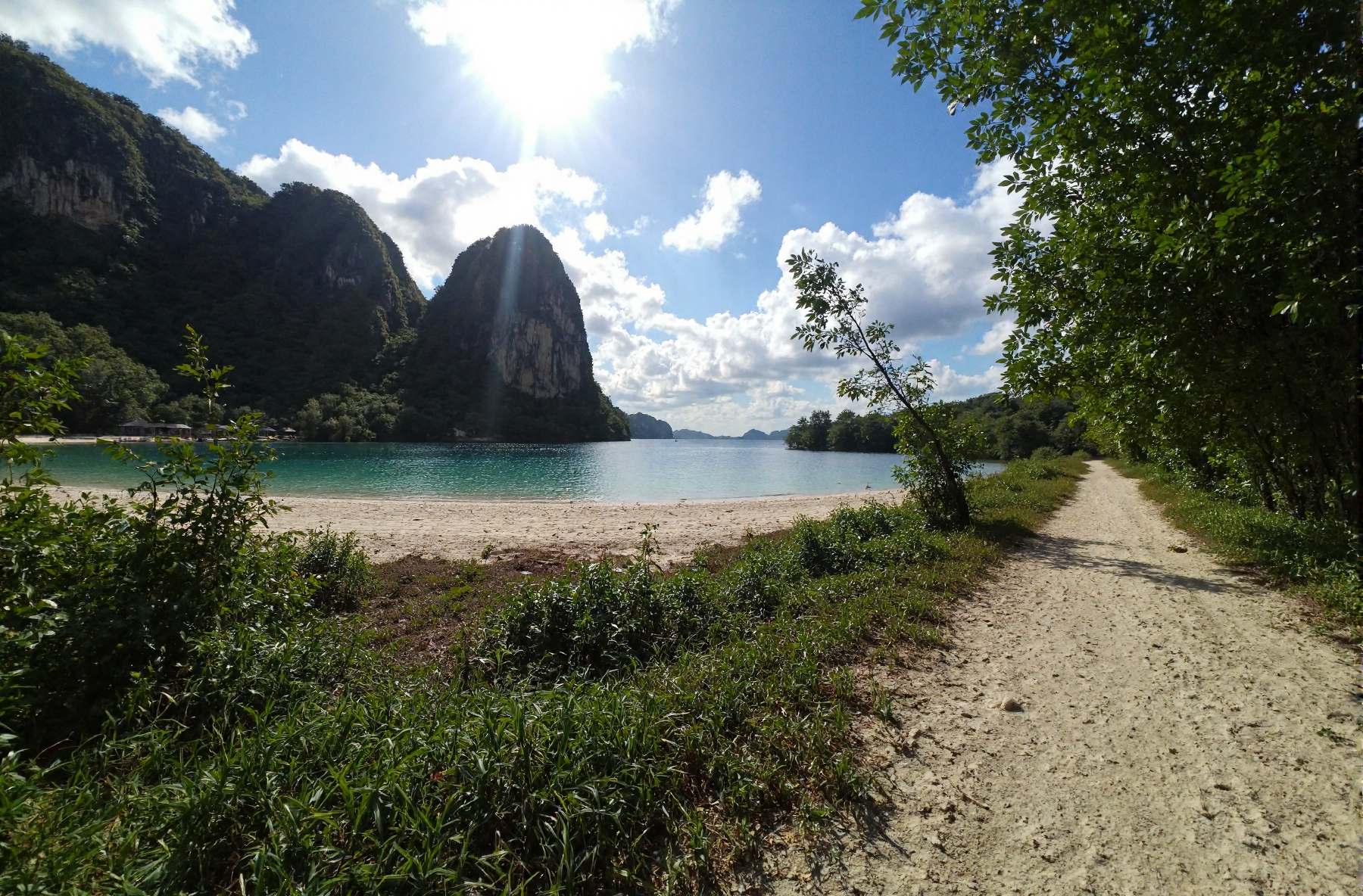 l3n0v0. ultra-wide pov, from ground view angle, lush tropical archipelago, towering jungle-covered cliffs and dramatic rocky outcrop dropping into a turquoise lagoon, ivory crescent beache along the shoreline, crystal-clear water reflecting cotton-candy cumulus clouds, sun-bleached dirt path snaking through dense emerald foliage, distant bamboo huts barely visible through the canopy, intense high-noon sunlight casting dramatic bloom and lens flare over the scene, deep shadows enveloping the foreground for contrast, subtle heat shimmer rising from treetops, gentle coastal breeze swaying palm crowns, early-2000s digital camera aesthetic with soft sensor grain and chromatic edges. amateur quality, candid style