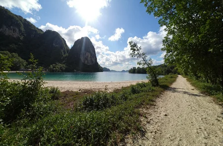 l3n0v0. ultra-wide pov, from ground view angle, lush tropical archipelago, towering jungle-covered cliffs and dramatic rocky outcrop dropping into a turquoise lagoon, ivory crescent beache along the shoreline, crystal-clear water reflecting cotton-candy cumulus clouds, sun-bleached dirt path snaking through dense emerald foliage, distant bamboo huts barely visible through the canopy, intense high-noon sunlight casting dramatic bloom and lens flare over the scene, deep shadows enveloping the foreground for contrast, subtle heat shimmer rising from treetops, gentle coastal breeze swaying palm crowns, early-2000s digital camera aesthetic with soft sensor grain and chromatic edges. amateur quality, candid style