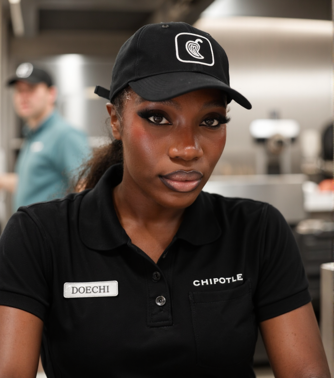 close-up headshot portrait shot on Fujifilm XT-50 of doech11 black woman working behind the counter at a Chipotle Mexican Grill resturaunt <lora:flux_realism_lora:.75> <lora:Hand v2:1> doechii, the woman has plump lips with shiny lipgloss, the woman has a seductive look on her face, young adult woman in her mid 20's, looking at the viewer and making eye contact with the viewer, beautiful dark-skinned woman, pursed plump DSLs, <lora:doechii:1> mouth closed, seductive gaze, the woman is wearing a black polo with a silver nameplate above the left breastpocket with text on it that reads "DOECHII", the woman is wearing a black cap with the Chipotle logo on it, flirty happy woman, fast food kitchen background, shallow depth of field blurred background