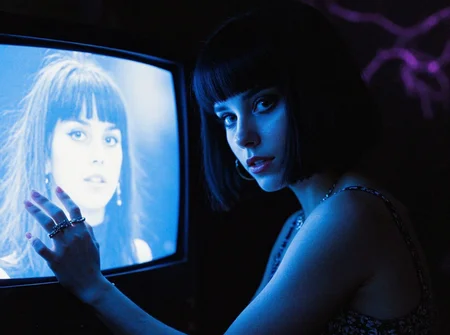 8n8log, Young woman, late 20s, sleek black hair with blunt bangs, wearing a patterned sleeveless top, dark eyeliner, glossy lipstick, multiple silver rings and ear piercings, poised with her left hand touching an old television screen displaying her face in monochrome, ambient blue lighting, shadows adding depth to her features, the background shows hints of purple light resembling a storm, image displays a surreal atmosphere, slightly blurred details, high contrast. film photography aesthetic, film grain effect prominent throughout image, high contrast lighting creating dramatic shadows, grainy film-like texture, professional photography technique, dramatic chiaroscuro lighting effect