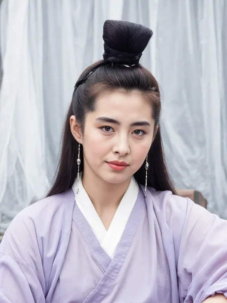 This is a high-resolution photograph of a young Asian woman with a fair complexion, styled in traditional Korean attire. She has long, straight black hair, adorned with a small, intricately braided bun at the top, and wears a pale lavender hanbok with a white inner robe. Her makeup is minimal, highlighting her natural beauty. She gazes directly into the camera with a calm expression, framed by delicate, dangling earrings. The background features soft, flowing white drapes, creating a serene, ethereal atmosphere. The lighting is soft and diffused, enhancing her features and the fabric's texture. The image follows the rule of thirds, with the woman's face and upper body positioned in the upper third, drawing attention to her serene expression and traditional attire. The overall composition exudes elegance and tranquility.
