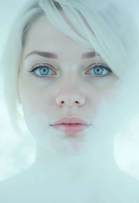 A closeup, surreal, ethereal portrait of a woman with a hauntingly beautiful, almost angelic presence. Her pale complexion is luminescent, blending seamlessly into the pure white background, creating an impression that she is emerging from light itself. Her very large piercing blue eyes are the focal point, their crystalline clarity and soft glow conveying a sense of otherworldly wisdom and calm. Her long lashes are bold and dramatic, contrasting starkly against her porcelain skin, drawing the viewer's gaze deeper into her haunting stare. Her features are delicately sculpted, with high cheekbones, a perfectly straight nose, and soft, full lips that hold a subtle, neutral expression, suggesting serenity and quiet strength. Her platinum-white hair flows and floats softly upwards, emphasizing her celestial appearance. The fine strands are light and wispy, blending seamlessly into the white backdrop with white ashes falling around her, adding to the ethereal atmosphere. The lighting is diffused and even, erasing shadows and creating a minimalistic aesthetic that places full focus on her striking features. The overall composition feels timeless, as if capturing the essence of purity and perfection, with a soft, dreamlike quality that invites introspection and awe. The simplicity of the image enhances its mystique, creating a profound, angelic presence that feels both familiar and otherworldly. <lora:MysticFantasy:0.2318463134765626>