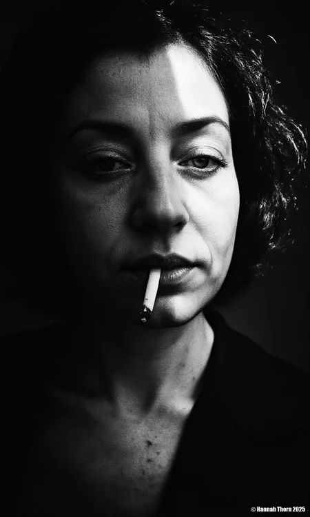 A close-up, black and white portrait in the vein of Henri Cartier-Bresson, channeling the mystery of film noir. A woman, captured with intense, shadowed eyes like a Diane Arbus subject, stares directly at the viewer, a half-smoked cigarette dangling from her lips, casting a Rembrandt-esque shadow across the texture of her skin. The stark monochrome palette, reminiscent of Man Ray, emphasizes the details of her face, creating a sense of raw intimacy and unwavering confidence. The image, with its dramatic shadows and high contrast, evokes the cinematic style of the 1940s<lora:_Flux\Anders_Petersen.safetensors:1.0:1.0>