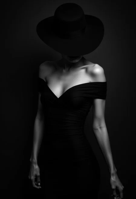 in a dimly lit, a monochrome high-fashion portrait of a woman donning an exquisite black dress with matching wide-brimmed hat against a darkened, blurred background. Her fitted, off-the-shoulder gown wraps around her bodice, cinched at the waist by a matching sash. The hat's shadow obscures her face, infusing mystery into the scene. Dark shot, dramatic shadows, muted highlights, black over black, chiaroscuro, film noir style.