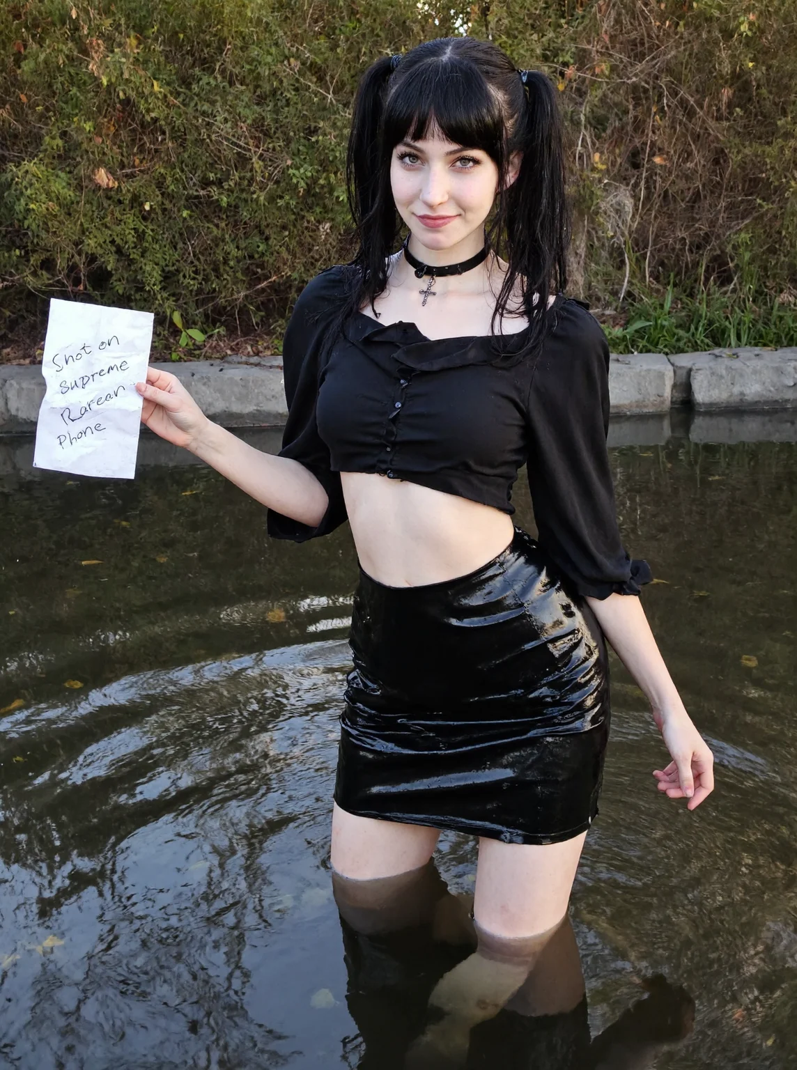 s2ms8ng, visible sensor noise, artificial over-sharpening, heavy HDR glow. A young woman in her early 20s, light skin, eastern european facial features, dark gothic pigtails with bangs. She stands awkwardly in a pond in a city park. her legs are submeged in water. Amateur photo, blown-out highlights and crushed shadows. She wears a black choker and a cross necklace, black loose crop top and black pvc wet pencil skirt, gothic vibe. Expression: sarcastic smirk, looking at the viewer. In her left hand, she holds a wrinkled piece of paper with the scribbled text "Shot on Supreme Korean Phone". Background: grainy bushes, reflective water with light artifacts. Daytime