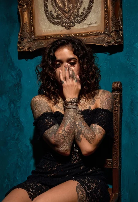 The image is a high-resolution photograph of a young woman with a light to medium skin tone and curly, dark brown hair styled in loose, voluminous waves. She is seated on a chair with her legs spread wide, positioned against a textured, teal-blue wall adorned with two framed, gold-leafed artworks. One of the framed artworks shows a detailed, stylized portrait, and the other is a more abstract piece. The woman is wearing a sheer, black, off-the-shoulder dress with intricate lace detailing, which drapes loosely around her body, revealing her arms and upper chest.
Her arms are heavily tattooed with intricate designs, including a prominent tattoo of a rose on her left arm. She is covering her face with her hands, obscuring her features, adding an air of mystery. She wears several layers of layered, delicate necklaces and bracelets, including a prominent, ornate cross necklace and a chunky, silver bracelet. The lighting is dramatic, casting soft shadows and highlights that accentuate the textures and contours of her skin and the intricate patterns on her dress and tattoos. The background's textured wall and the wooden chair with ornate, wrought iron detailing provide a rich, artistic ambiance. The overall mood is a blend of edgy, artistic, and slightly melancholic.
