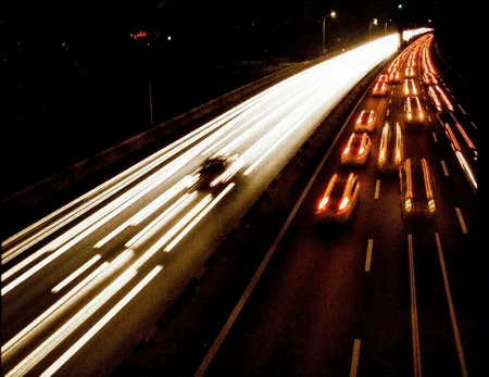 8n8log, view from the top of a bridge overlooking a highway at night. The highway is filled with cars traveling at high speed, represented mainly by the light trails of their rear and front lights, creating a dynamic, blurry effect. In the center of the scene, nestled among these light trails of cars.
film photography aesthetic, film grain effect prominent throughout image, high contrast lighting creating dramatic shadows, grainy film-like texture, dramatic chiaroscuro lighting effect, amateurish quality, candid style