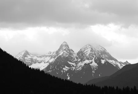 Snow-capped mountain range, rugged peaks, dark jagged edges, overcast sky, thick cloud cover, dramatic sky and clouds, dense forest at mountain base, black and white image, high contrast, shadowed foreground, slight haze in atmosphere, outdoor natural landscape, diffuse lighting conditions, monochrome., 