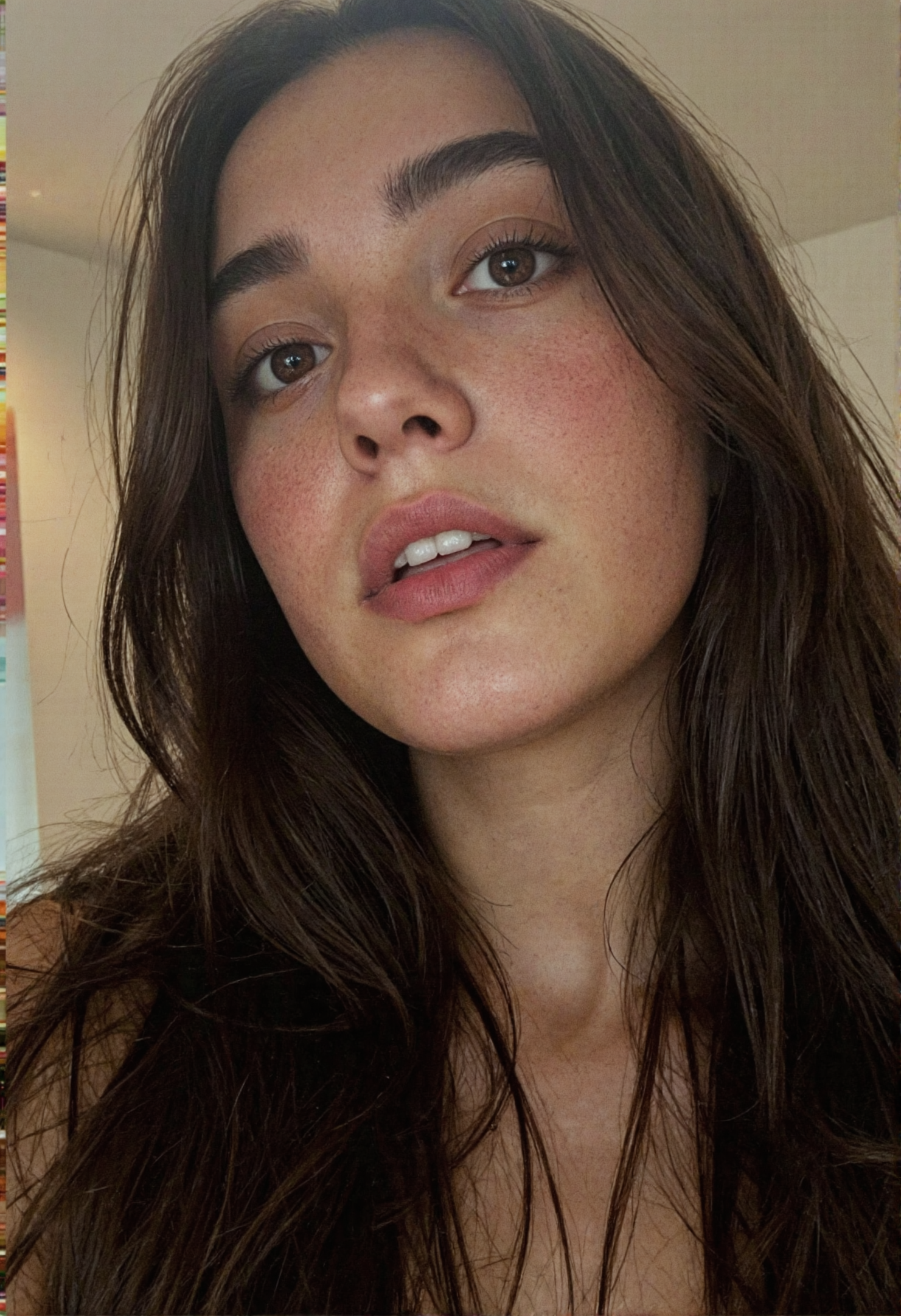 A tightly composed, extreme close-up photograph of a lovely woman's face, captured from a slightly low angle to heighten a sense of presence and intimacy. The top of the frame trims just above her gently arched eyebrows, while the bottom edge cuts off below her full, slightly parted lips — revealing the upper edges of her clean, white teeth and the faintest glimpse of her tongue. Her eyes, a soft amber-brown, are the focal point: luminous and glassy, with intricate iris striations and the subtle moisture that clings to the lower lid, catching the light. Her lashes are long, dark, and cleanly defined, curling softly above and below her gaze, enhancing the natural shape of her eyelids.

Her skin is smooth and even-toned, with a realistic, natural texture that glows under soft, diffuse lighting. A faint flush warms her upper cheeks, hinting at youth and vitality. Wisps of rich, dark hair frame the sides of her face, catching a bit of reflected light and hinting at a deep black or espresso tone. The lighting is frontal and gentle, eliminating harsh shadows while preserving micro-details like skin texture and lash separation. The background is a pale, nearly white gradient — minimal and clean — drawing full attention to her face without distraction. The image feels breath-close, intimate, and reverent, capturing subtle expressions and the delicate balance of tension and calm in her features.
