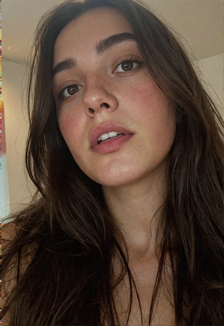 A tightly composed, extreme close-up photograph of a lovely woman's face, captured from a slightly low angle to heighten a sense of presence and intimacy. The top of the frame trims just above her gently arched eyebrows, while the bottom edge cuts off below her full, slightly parted lips — revealing the upper edges of her clean, white teeth and the faintest glimpse of her tongue. Her eyes, a soft amber-brown, are the focal point: luminous and glassy, with intricate iris striations and the subtle moisture that clings to the lower lid, catching the light. Her lashes are long, dark, and cleanly defined, curling softly above and below her gaze, enhancing the natural shape of her eyelids.
Her skin is smooth and even-toned, with a realistic, natural texture that glows under soft, diffuse lighting. A faint flush warms her upper cheeks, hinting at youth and vitality. Wisps of rich, dark hair frame the sides of her face, catching a bit of reflected light and hinting at a deep black or espresso tone. The lighting is frontal and gentle, eliminating harsh shadows while preserving micro-details like skin texture and lash separation. The background is a pale, nearly white gradient — minimal and clean — drawing full attention to her face without distraction. The image feels breath-close, intimate, and reverent, capturing subtle expressions and the delicate balance of tension and calm in her features.