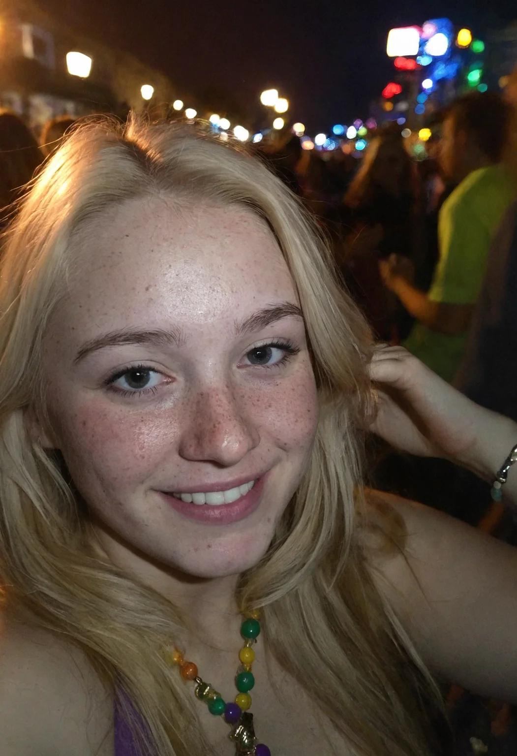 Amateur cellphone photography capturing a cute and attractive young woman with blonde hair at Mardi Gras during a stunning sunset. The scene is vibrant with the festive atmosphere of the parade, colorful beads, and lively crowds in the background. The woman has a playful smirk on her face, exuding confidence and charm, with subtle freckles (freckles:0.2) adding a touch of natural beauty. The photo is taken with a Samsung Galaxy in challenging lighting conditions—poor lighting, low light, and underexposed—resulting in a high-contrast image with deep shadows and bright highlights. The composition emphasizes the woman as the focal point, with the warm hues of the sunset casting a golden glow on her features, while the surrounding environment remains moody and atmospheric. The imperfections in the photo, such as graininess and slight blur, add to the raw, authentic feel of amateur photography.