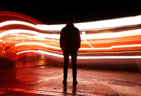 Lines of Movement, A night-time urban scene featuring a lone figure standing still in the center of the frame, with vibrant red, orange, and white light trails swirling around them, captured with long exposure to create a sense of motion and energy. The figure, dressed casually in a jacket and jeans, is illuminated by the surrounding light, their silhouette contrasting against the dark night. The ground beneath them is wet, reflecting the bright lights and adding depth to the scene. The light trails, possibly from passing cars or other fast-moving vehicles, streak across the image in curved lines, wrapping around the figure and adding a surreal, almost futuristic feel to the composition. The lighting is moody, with a rich contrast between the dark background and the bright, colorful light streaks. The overall atmosphere is one of stillness amidst chaos, blending human presence with the fast-paced movement of the city.