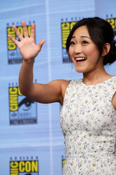 karenfukuhara, 1girl, brown eyes, standing, photo(medium), on stage, comic-con background, white floral dress, looking at someone, shocked expression, waving to someone,  cinematic lighting, photorealistic, ultra-sharp, detailed, professional photographer, black hair, blurry background, bokeh,  rule of thirds