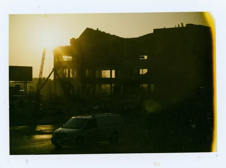 p0l2rd, Silhouette of a partially demolished building in the foreground, illuminated by the golden glow of the setting sun. The structure is surrounded by a haze of dust, with beams of sunlight piercing through. Visible cranes and machinery contribute to the industrial scene. The street below shows a billboard, an ambulance, and a few scattered vehicles. The sky is clear with faint contrails, underexposed. prominent film grain, high contrast lighting casting dramatic shadows, grainy film-like texture, polaroid-style format with white border, distinctive burnt edge, photograph appears aged or partially developed, with the right side fading into white